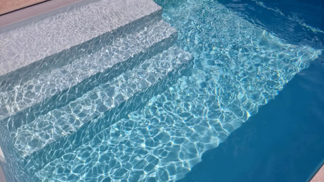 Treppenstufen, die in einen klaren blauen Pool führen, mit Sonnenlicht, das auf der Wasseroberfläche reflektiert wird.
