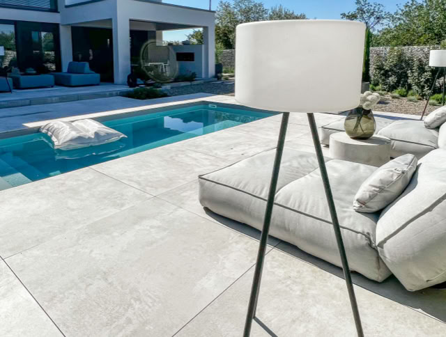 Moderne Terrasse mit Pool, Liegestühlen und Stehlampe, im Hintergrund ein Haus.
