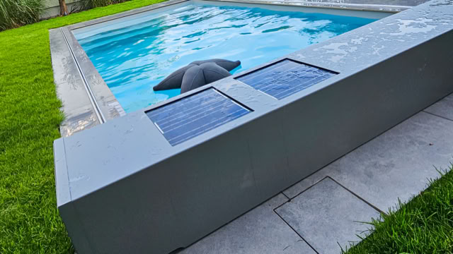 Ein rechteckiger Pool mit blauen Fliesen, umgeben von grünem Rasen und grauen Steinplatten. Ein schwarzer Sonnenschirm ist teilweise im Wasser.
