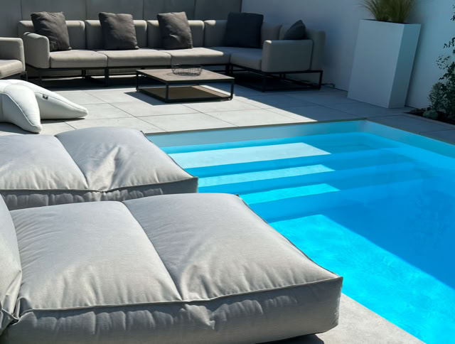 Sonnenliegen und ein Sofa mit Kissen neben einem blauen Pool auf einer Terrasse.