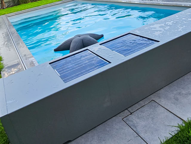 Ein rechteckiger Pool mit blauen Fliesen, umgeben von grünem Rasen und grauen Steinplatten. Ein schwarzer Sonnenschirm ist teilweise im Wasser.