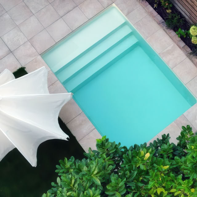 Kleiner rechteckiger Pool mit blauen Wasser, umgeben von Fliesen, einem weißen Sonnenschirm und grünen Pflanzen.