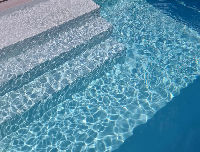 Treppenstufen, die in einen klaren blauen Pool führen, mit Sonnenlicht, das auf der Wasseroberfläche reflektiert wird.